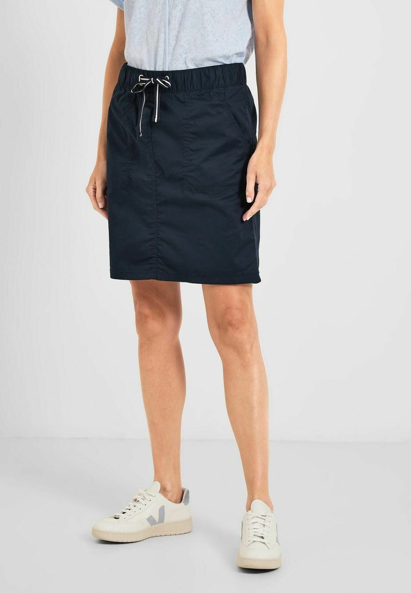 Falda azul marino, hasta la rodilla, con cintura elástica, cordón y bolsillos laterales, combinada con zapatillas de color claro.