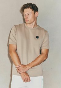 Camiseta de manga corta beige con cuello redondo, que presenta un pequeño parche con logotipo negro en el pecho y una tela suave y texturizada.