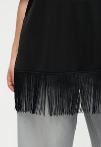 Μαύρο τοπ με μακριά φούντα στο τελείωμα που φοριέται πάνω από ανοιχτό γκρι παντελόνι, με μερική θέα του αριστερού χεριού και μπράτσου σε απλό φόντο.