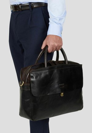 Mann iført blå skjorte og marineblå bukser holder en svart lærveske i håndtakene ved siden av seg.