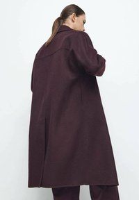 Weinroter Wollmantel mit langem, locker geschnittenem Design, einem einzelnen Rückenschlitz und einem Reverskragen. Die Textur wirkt weich und warm.