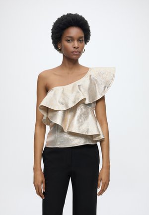 Γυναίκα με κοντά κατσαρά μαλλιά, που φορά χρυσή μπλούζα με έναν ώμο και βολάν και μαύρο παντελόνι, κοιτάζοντας μπροστά σε απλό λευκό φόντο.