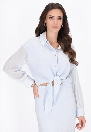 Kvinde med langt bølget hår iført lyseblå knappet bluse bundet i taljen og matchende nederdel, poserer med den ene hånd på hoften.