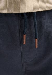 Sweatshirt en mélange de coton beige à texture côtelée, associé à un pantalon bleu foncé avec cordons de serrage noirs et embouts métalliques bronze.