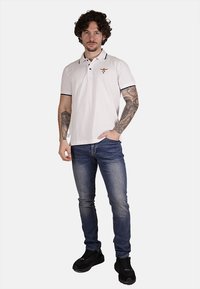 Maglietta polo bianca con accenti neri sul colletto e logo, abbinata a jeans denim scuri e sneakers nere. Il modello ha capelli ricci e tatuaggi.