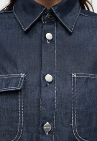 Σκούρο μπλε πουκάμισο από τζιν ύφασμα με κουμπιά μπροστά, μυτερό γιακά και τσέπες στο στήθος, διακοσμημένο με μεταλλικά κουμπιά και ραφές αντίθετου χρώματος.