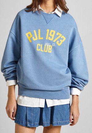 Sweatshirt in cotone blu con grafica gialla testuale "PJL 1973 CLUB" e "PHYS. ED." sotto. Presenta polsini e orlo a costine, indossato sopra una camicia bianca con colletto.
