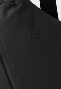 Μαύρη υφασμάτινη τσάντα ώμου με ανάγλυφο λογότυπο "BOSS" και ρυθμιζόμενο λουρί σε κοντινή λήψη.