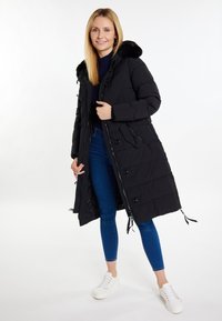 Sort pufferjakke med en faux fur-hætte, der har lynlås foran, knapper, sidelommer og et langt, quiltet design. Hvide sneakers på.