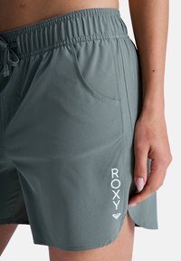 Ανοιχτό πράσινο μαγιό με ελαστική μέση, μπροστινές τσέπες και λευκό λογότυπο "ROXY" στην αριστερή πλευρά. Ομαλή υφή, casual σχεδιασμός.