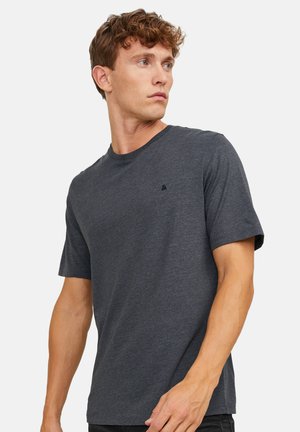 Jonge man met krullend haar, gekleed in een eenvoudige donkergrijze korte mouw t-shirt, kijkt naar links tegen een witte achtergrond.