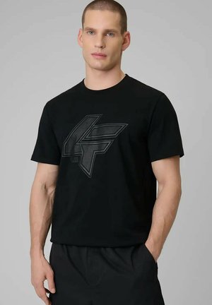 Man met kort haar die een zwart t-shirt draagt met een groot, gestructureerd geometrisch logo, staande met één hand in de zak tegen een effen grijze achtergrond.