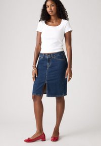 Gonna di jeans con spacco frontale, presenta un design a cinque tasche e una chiusura a bottone. Abbinata a un top aderente bianco e a delle ballerine rosse.
