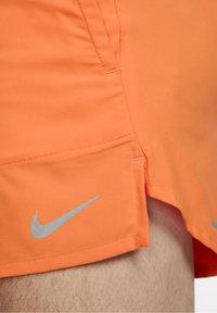 Nike urheilusortsit kirkkaan oranssina, joissa on teksturoitu kangas, sivuhaalari ja heijastava hopeinen logo alareunassa.