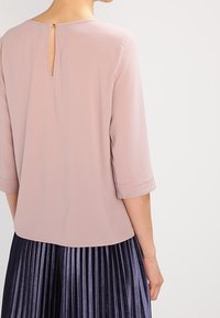 Donna che indossa un top rosa pallido con maniche a tre quarti e un piccolo buchino sul retro con bottone, abbinato a una gonna plissettata scura.