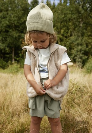Beige Fleece-Weste mit Reißverschluss, über einem hellen Shirt mit Grafik getragen. Olivgrüne Shorts, Kind passt den Bund an, im Freien.
