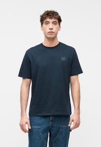 Νεαρός άνδρας με σγουρά μαλλιά φοράει ένα σκούρο μπλε μπλουζάκι και μπλε τζιν, στέκεται ευθεία μπροστά σε ένα απλό λευκό φόντο.