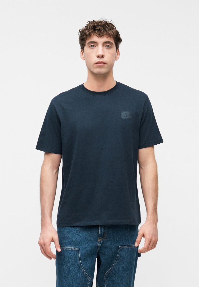 Νεαρός άνδρας με σγουρά μαλλιά φοράει ένα σκούρο μπλε μπλουζάκι και μπλε τζιν, στέκεται ευθεία μπροστά σε ένα απλό λευκό φόντο.