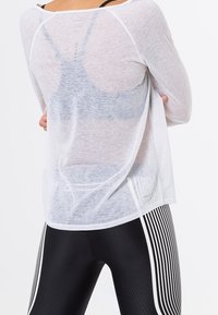 White, sheer long-sleeve top with a relaxed fit, revealing a black sports bra underneath. Paired with black leggings featuring vertical white stripes.