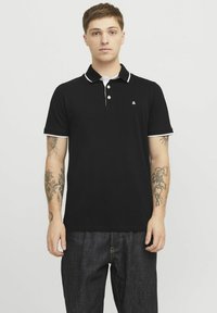 Zwarte poloshirt met witte kraag en manchetaccenten, voorzien van drie knoppen en een klein logo op de borst. Draagt met donkere denim broek.