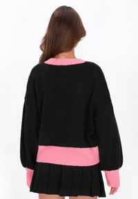 Vrouw met lang bruin haar draagt een zwarte trui met roze manchetten en zoom, gecombineerd met een geplooide zwarte rok, met de rug naar de kijker gekeerd.