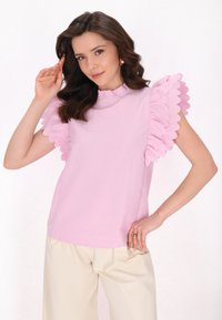 Rosa Oberteil mit gewelltem Rüschensaum, rundem Ausschnitt, strukturiertem Stoff; kombiniert mit hellfarbigen, lockeren Hosen. Einfaches, sauberes Design.