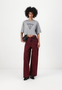 T-shirt grigia tagliata con logo nero, abbinata a jeans borgogna a gamba larga. La modella tiene una piccola borsa nera e indossa scarpe nere.