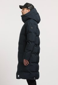 Tumesinine puffjakkk koos kõrge kraega ja kapuutsiga, millel on suur logo plaastriga varrukal ning pikem disain lisasoojustamiseks.