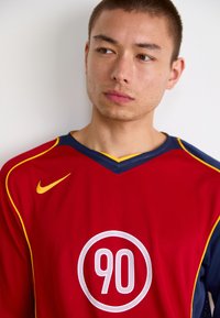 Rød og marineblå fodboldtrøje med et gult Nike-logo og en cirkulær hvid grafik med "90" i midten.