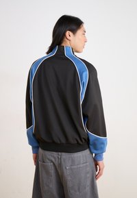 Black jacket with blue and white accents, featuring a high collar and textured fabric. The design includes a ribbed waistband and cuffs.