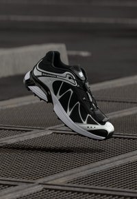 Czarno-biała biegałówka z siateczkową cholewką, zakrzywioną podeszwą i zintegrowanym systemem sznurowania, prezentująca dynamiczny, aerodynamiczny design.