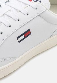 Biały skórzany sneaker z płaskimi sznurowadłami, z logo w kolorach czerwonym, białym i niebieskim. Podeszwa o strukturze z jasną krawędzią.