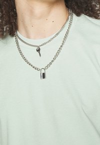 Stříbrné řetízkové náhrdelníky vrstvené, s přívěskem ve tvaru klíče a malým zámkem. Nosené přes světle zelenou strukturovanou košili.