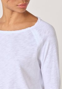 Frau trägt ein weißes Langarmshirt mit einem rohen, abgeschnittenen Ausschnitt, der einen Teil ihres Halses und ihres unteren Gesichts vor einem einfarbigen Hintergrund zeigt.