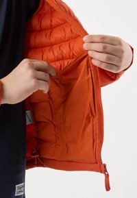 Mains tenant l'intérieur ouvert d'une veste matelassée orange vif montrant une poche intérieure et une doublure en tissu.