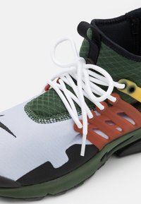 Zielono-biały but sportowy z białymi sznurowadłami, czarno-pomarańczowymi detalami w postaci klatki oraz czarną podeszwą, widoczny z boku pod kątem.