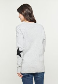 Pulover tricotat de culoare gri deschis, cu un design de stea neagră pe mâneca stângă, având o croială lejeră și manșete și margine ribbed.