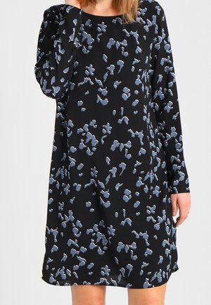 Mujer con un vestido negro de manga larga, de corte holgado, con un estampado floral abstracto disperso en azul y blanco.