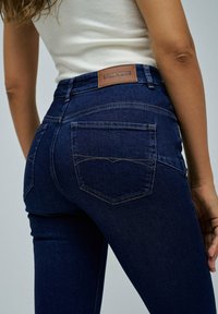 Jeans de mezclilla azul oscuro con un diseño ajustado, que cuentan con un parche de cuero en la cinturilla y dos bolsillos traseros con detalles de costura.
