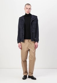 Marineblauwe jas met meerdere voorzakken, gedragen over een zwarte coltrui, gecombineerd met beige broek en zwarte schoenen. Soepele stof en een getailleerde pasvorm.