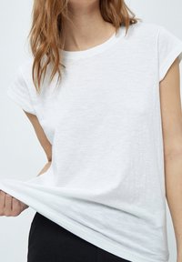 T-shirt blanc à manches courtes en tissu léger légèrement texturé. Encolure ronde, coupe décontractée. Design simple sans ornements.