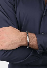 Bracciale intrecciato nero con accenti in metallo argentato, dotato di una chiusura cilindrica. Indossato su un polso con una camicia blu navy.