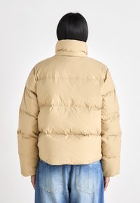 Giacca puffer beige con colletto alto, caratterizzata da una texture trapuntata e un design voluminoso. Indossata sopra jeans in denim azzurro chiaro.