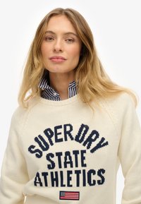 Femme blonde portant un pull crème avec le texte "SUPERDRY STATE ATHLETICS" et un petit patch de drapeau américain, ainsi qu'une chemise à col rayé bleu.