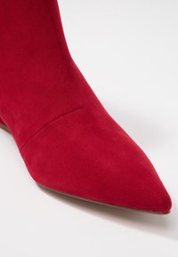 Czerwone zamszowe botki za kostkę z wąskim noskiem i gładką powierzchnią. Posiadają minimalne przeszycia na cholewce oraz smukły obcas.