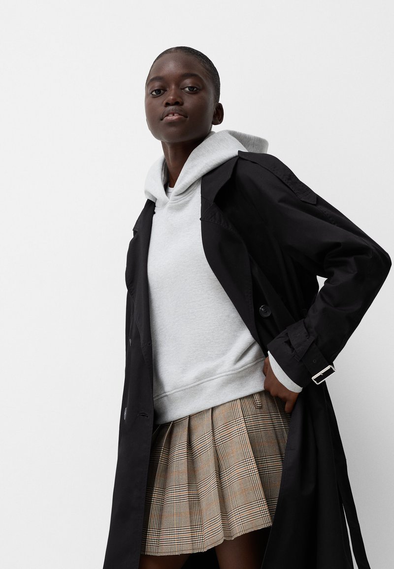 Młoda kobieta ubrana w szaro-skalny kaptur, czarny płaszcz i plisowaną spódnicę w kratę na jednolitym białym tle.