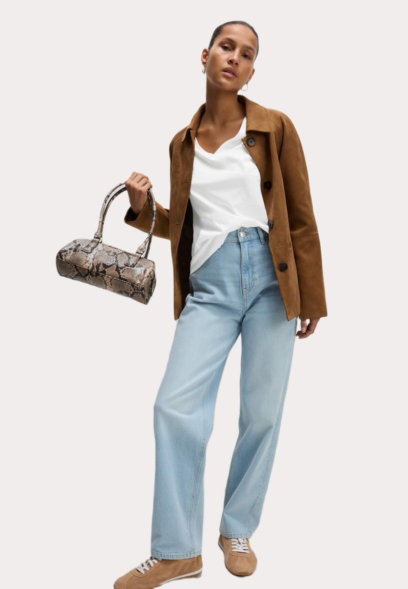 Giacca in suede marrone, maglietta bianca a scollo a V, jeans a gamba larga blu chiaro e sneakers beige. Tiene una borsa piccola con motivo a serpente.