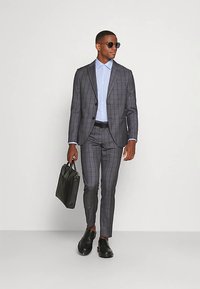 Grijs geruit pak met een op maat gemaakte jas en smalle broek, gecombineerd met een lichtblauw overhemd. Een zwarte aktetas en een zonnebril maken de look compleet.