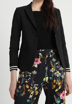 Mujer con una blazer negra con puños a rayas, una camiseta negra y pantalones negros con un estampado floral colorido.