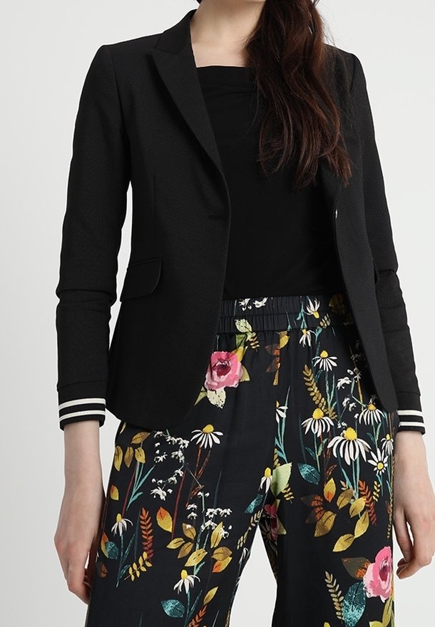 Femme portant un blazer noir avec des poignets rayés, un haut noir et un pantalon noir avec un motif floral coloré.